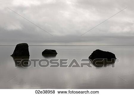 Stock Photo - Three solitary rocks rising from the calm ocean against the horizon on a cloudy day. Fotosearch