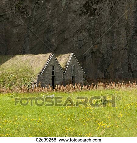 Stock Photo - Weathered dwellings with grass on roof under a mountain cliff in a meadow. Fotosearch