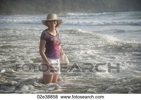 Stock Photo - Woman in a hat smiling standing in the ocean surf at knee height. Fotosearch