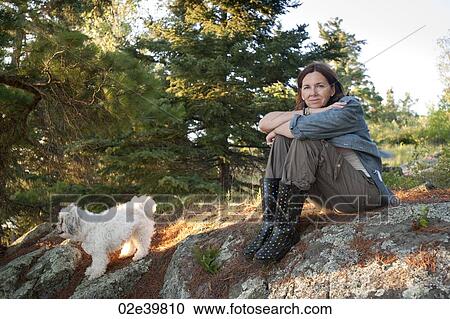 Stock Image - Woman sitting on a rock with her white dog at Lake of the Woods, Ontario. Fotosearch