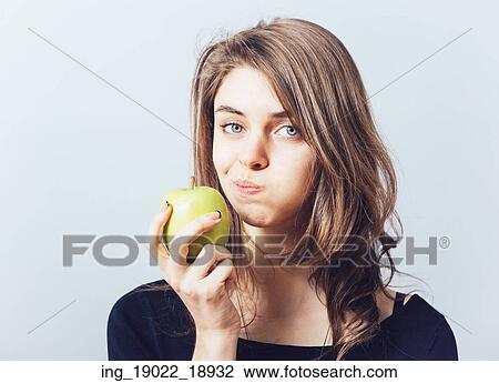 Stock Image - Beautiful young woman bites a green apple on a gray background. Fotosearch