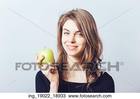 Stock Image - Beautiful young woman bites a green apple on a gray background. Fotosearch