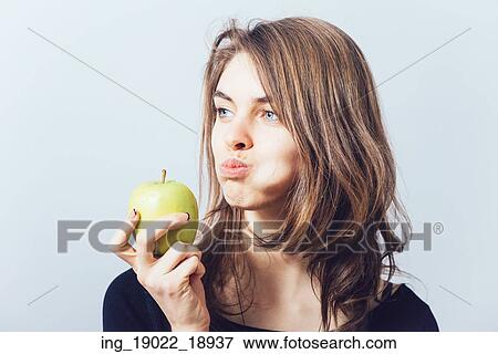 Stock Photo - Beautiful young woman bites a green apple on a gray background. Fotosearch