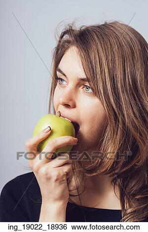 Beautiful young woman bites a green apple on a gray background View Large Photo Image Stock Photograph - Beautiful young woman bites a green apple on a gray background. Fotosearch
