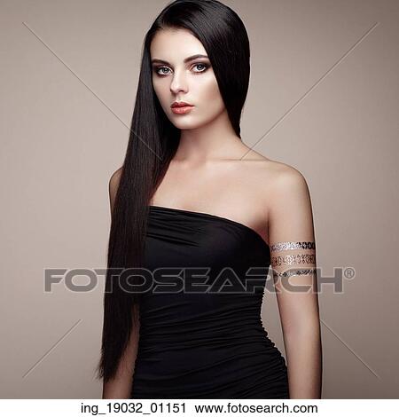 Stock Image - Fashion portrait of elegant woman with magnificent hair. Brunette girl. Perfect make-up. Girl in elegant dress. Flash tattoo gold. Fotosearch