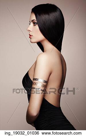 Stock Photograph - Fashion portrait of elegant woman with magnificent hair. Brunette girl. Perfect make-up. Girl in elegant dress. Flash tattoo gold. Fotosearch