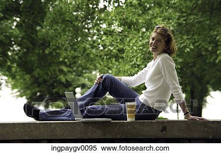 Young woman sitting on a ledge in a garden with a laptop computer View Large Photo Image Stock Photography - Young woman sitting on a ledge in a garden with a laptop computer. Fotosearch