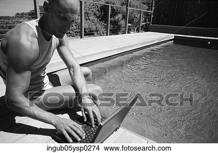 Picture - Close-up of a young man sitting at a poolside with a laptop. Fotosearch