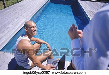 Stock Photo - High angle view of a young man sitting at a poolside and talking to another person. Fotosearch