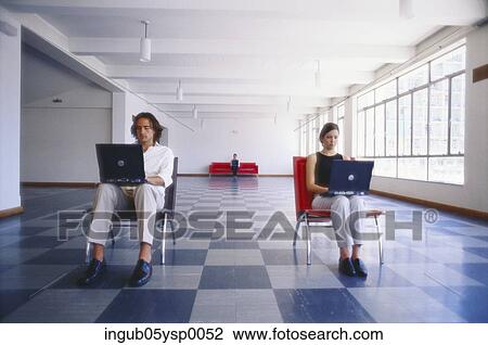Stock Image - Young man and young woman sitting on chairs and using laptops. Fotosearch