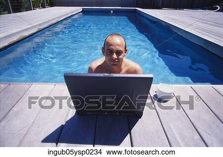 Picture - Young man in a swimming pool with a laptop on the ledge. Fotosearch