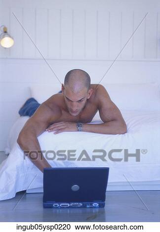 Stock Image - Young man lying on a bed and working on a Laptop on the floor. Fotosearch