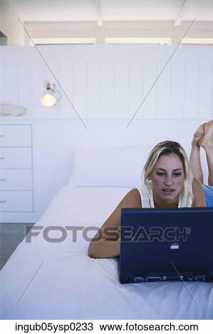 Stock Image - Young woman lying on a bed and working on a laptop. Fotosearch