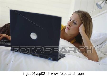 Stock Image - Young woman lying on a bed using a laptop. Fotosearch