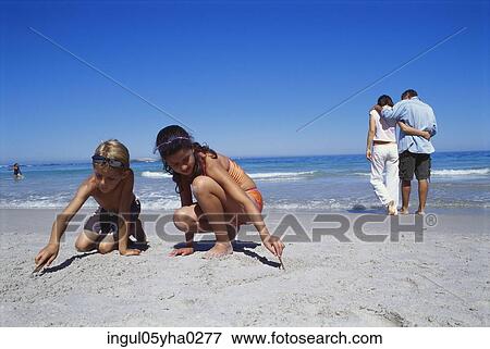 Girl and boy playing in the sand with their parents walking behind them View Large Photo Image Stock Photo - Girl and boy playing in the sand with their parents walking behind them. Fotosearch