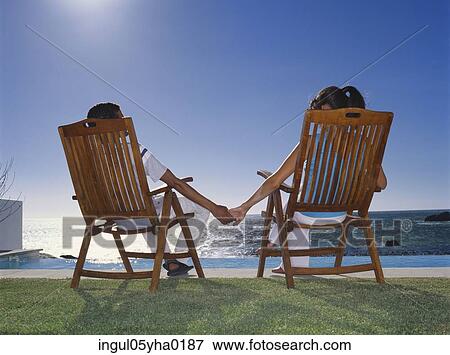 Rear view of a young couple sitting on a chair holding hands View Large Photo Image Stock Photo - Rear view of a young couple sitting on a chair holding hands. Fotosearch