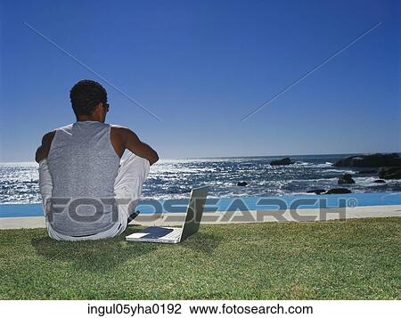 Stock Image - Rear view of a young man sitting on a lawn. Fotosearch