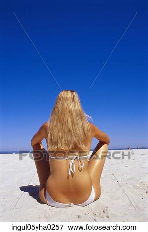 Stock Photo - Rear view of a young woman sitting on the beach. Fotosearch