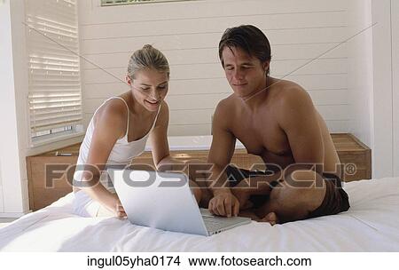 Picture - Young couple sitting on a bed working on a laptop. Fotosearch