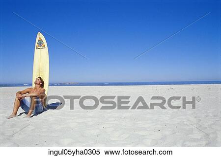 Stock Photography - Young man sitting on the beach in front of a surfboard. Fotosearch