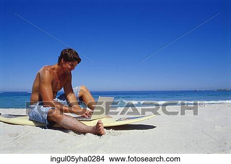 Picture - Young man sitting working on a laptop on the beach. Fotosearch