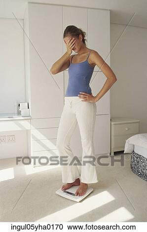 Stock Image - Young woman standing on a weighing scale covering her face. Fotosearch