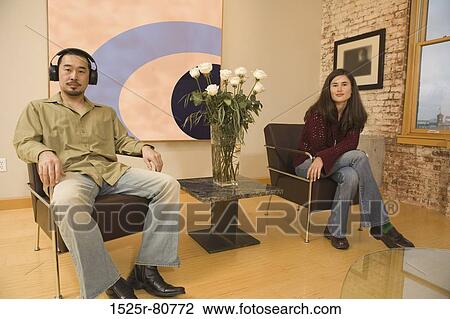 Stock Image - Portrait of a young man sitting on a chair wearing headphones with a young woman sitting beside him. Fotosearch