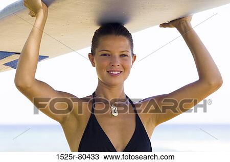 Stock Image - Portrait of a young woman carrying a surfboard. Fotosearch