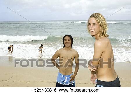 Stock Photograph - Two young men smiling on the beach with two young women standing in the background. Fotosearch