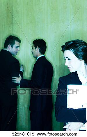 Stock Photography - Two business men talk behind the back of a business woman who is holding a laptop. Fotosearch
