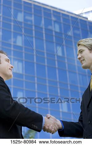 Two Businessmen stand together and give each other a handshake in front of a business building View Large Photo Image Picture - Two Businessmen stand together and give each other a handshake in front of a business building. Fotosearch