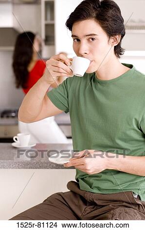 Portrait of a young man drinking tea in a kitchen with a young woman standing behind View Large Photo Image Picture - Portrait of a young man drinking tea in a kitchen with a young woman standing behind. Fotosearch