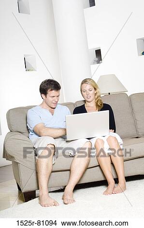 Picture - Young couple sitting on a couch and using a laptop. Fotosearch