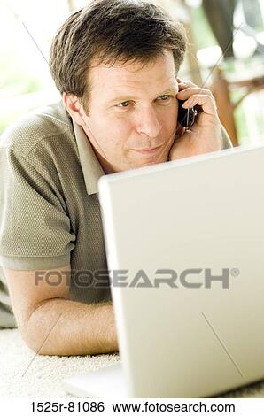 Stock Photograph - Young man lying in front of a laptop and talking on a mobile phone. Fotosearch