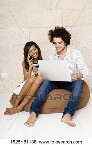 Young man sitting on a bean bag using a laptop with a young woman sitting beside him talking on a mobile phone View Large Photo Image Stock Photo - Young man sitting on a bean bag using a laptop with a young woman sitting beside him talking on a mobile phone. Fotosearch