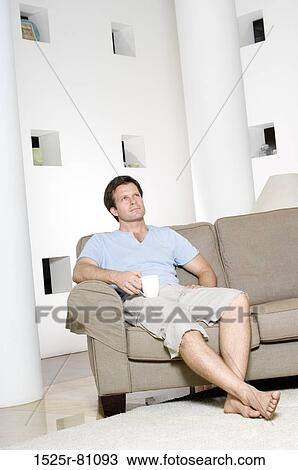 Young man sitting on a couch and holding a coffee cup View Large Photo Image Stock Image - Young man sitting on a couch and holding a coffee cup. Fotosearch