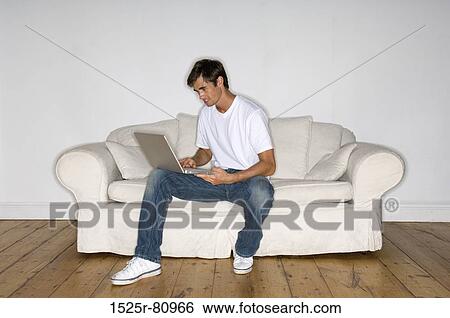 Young man sitting on a couch and using a laptop View Large Photo Image Stock Photograph - Young man sitting on a couch and using a laptop. Fotosearch