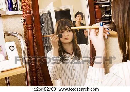 Stock Photo - Reflection of a teenage girl applying make-up to her face with her mother standing behind her. Fotosearch
