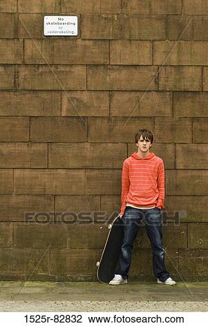 Teenage boy holding a skateboard standing in front of a stone wall View Large Photo Image Stock Image - Teenage boy holding a skateboard standing in front of a stone wall. Fotosearch