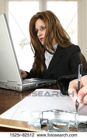 Stock Image - Businesswoman working on a laptop and writing on a clipboard. Fotosearch
