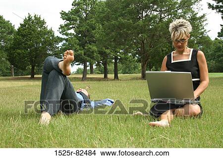 Mid adult woman working on a laptop with a man lying in the grass beside her View Large Photo Image Picture - Mid adult woman working on a laptop with a man lying in the grass beside her. Fotosearch