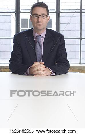 Businessman sitting at a table with his hands clasped in an office View Large Photo Image Stock Photography - Businessman sitting at a table with his hands clasped in an office. Fotosearch