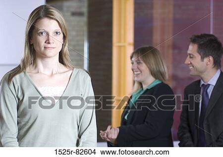 Picture - Businesswoman standing in an office with two business executives smiling in the background. Fotosearch
