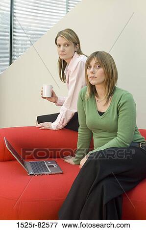 Mid adult woman sitting on a couch with another woman sitting behind her View Large Photo Image Stock Photo - Mid adult woman sitting on a couch with another woman sitting behind her. Fotosearch