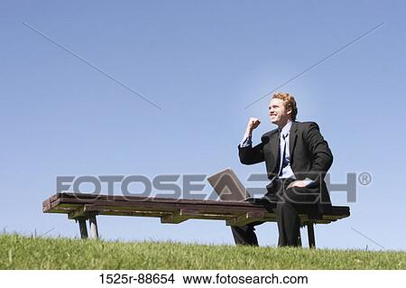 Business man in black suit and blue shirt and tie is holding one arm up in celebration, as he sits on a park bench, next to his laptop View Large Photo Image Picture - Business man in black suit and blue shirt and tie is holding one arm up in celebration, as he sits on a park bench, next to his laptop. Fotosearch