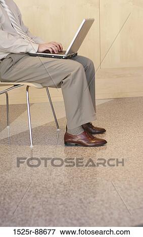 Stock Photo - Business man wearing tan shirt and brown shoes sits in a chair with his laptop in a business building. Fotosearch