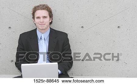 Stock Photography - Businessman in dark blue suit, blue shirt, and blue tie is sitting up against concrete wall and typing on his laptop. Fotosearch