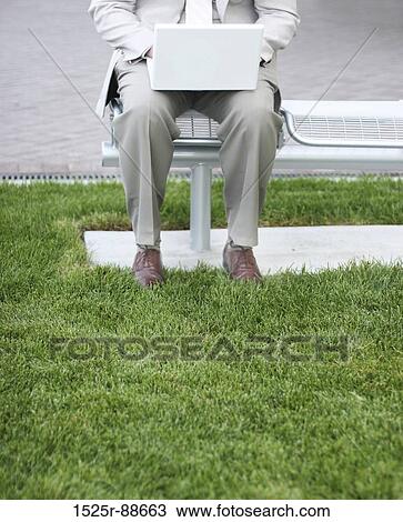 Businessman is sitting on metal bench next to the grass in his tan suit as he types on his laptop View Large Photo Image Stock Image - Businessman is sitting on metal bench next to the grass in his tan suit as he types on his laptop. Fotosearch