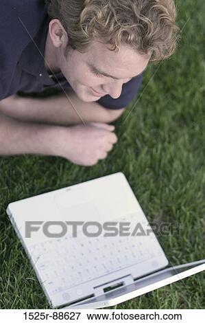 Happy young man dressed in blue shirt relaxes and lies down in the green grass, looking at his white laptop View Large Photo Image Stock Photo - Happy young man dressed in blue shirt relaxes and lies down in the green grass, looking at his white laptop. Fotosearch