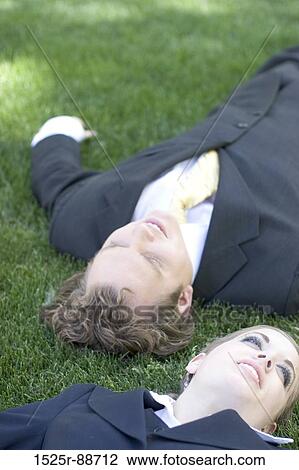 Stock Image - Two business people lying down in the grass in the park taking a break and smiling. Fotosearch